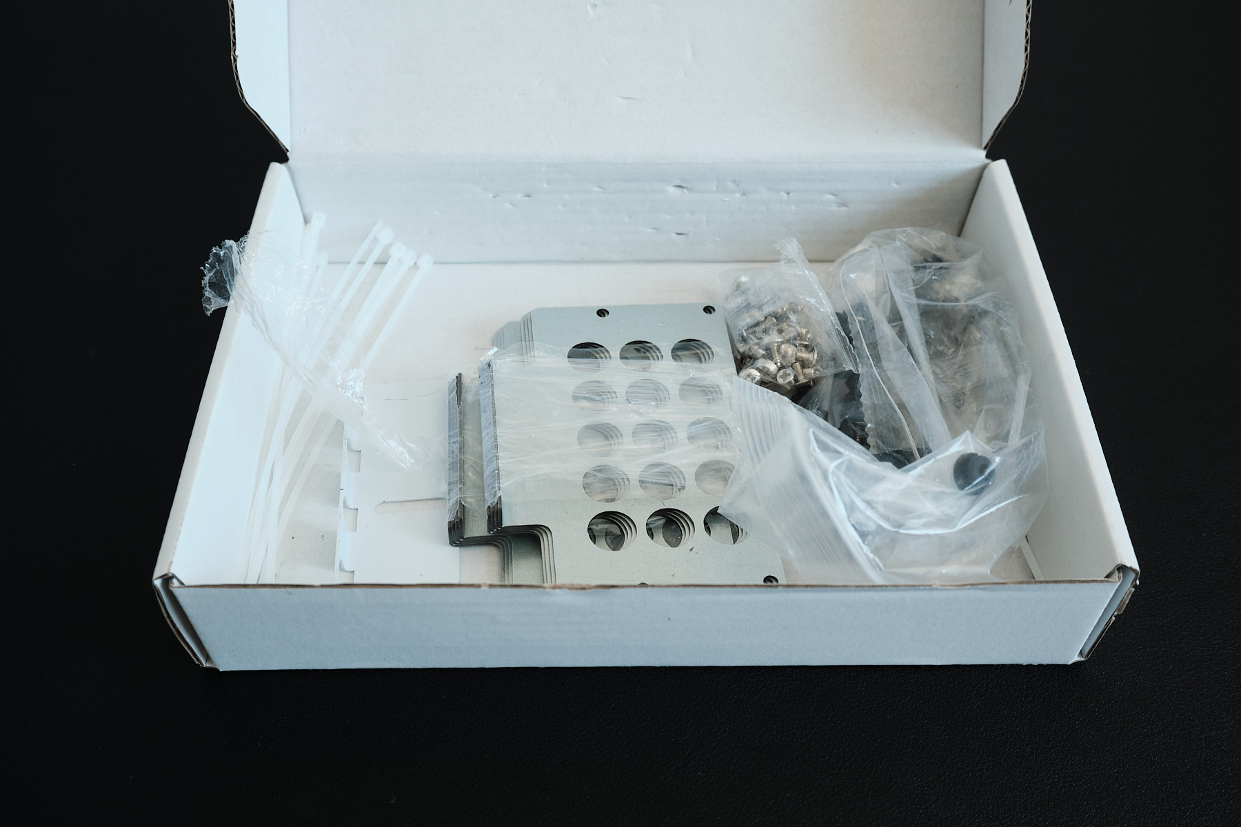 Open white accessory box containing metal hard drive mounting brackets, bags of screws, and cable ties