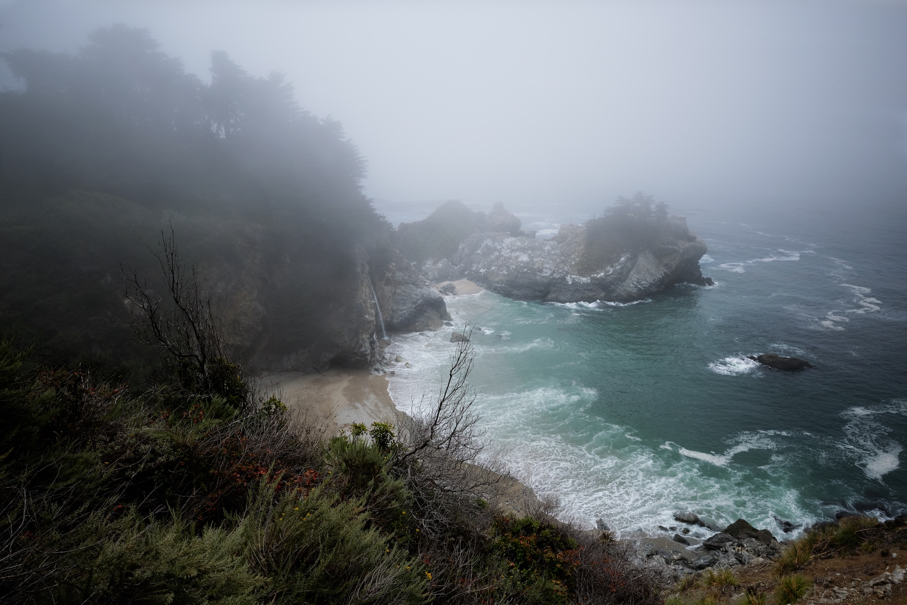 Foggy view of a secluded beach cove, turquoise waters, and rocky cliffs along a rugged coastline.
