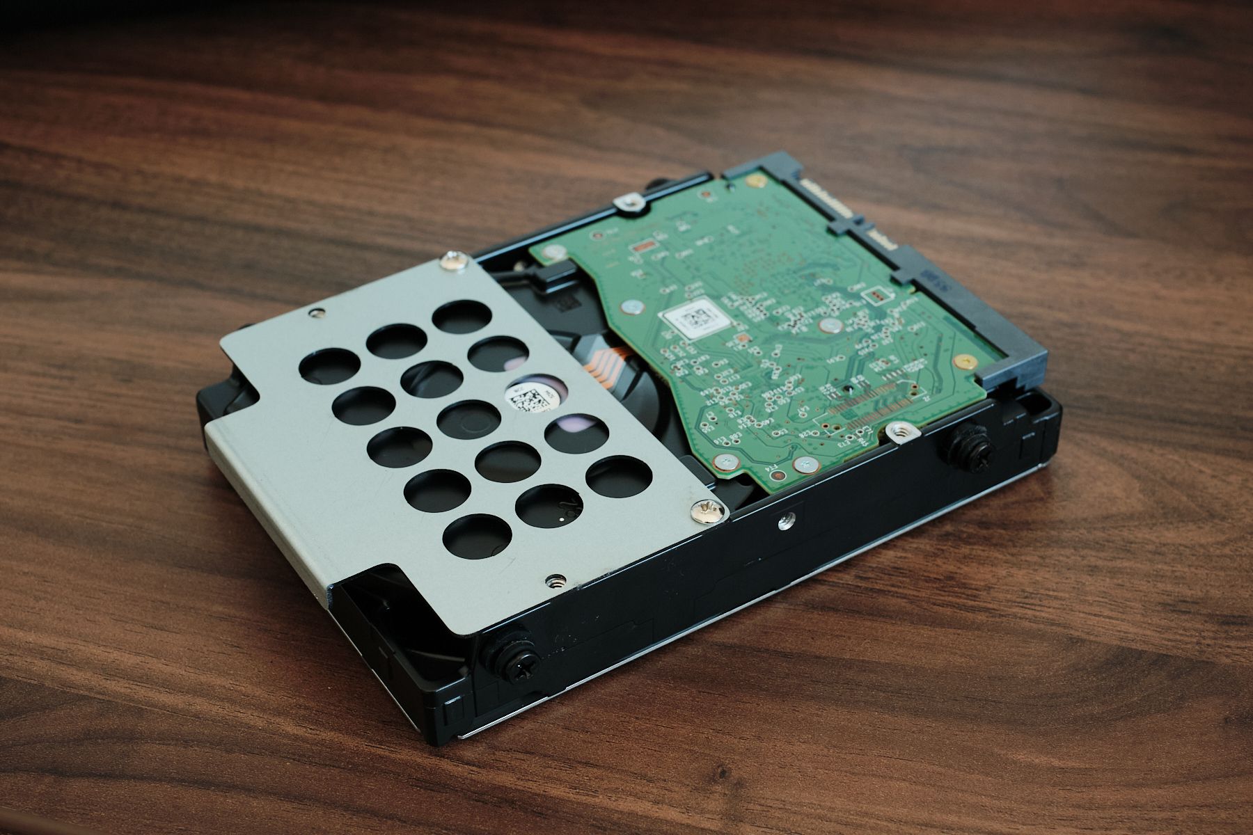 A 3.5-inch hard drive lying upside down on a wooden desk, with a perforated metal mounting tab screwed to one end and rubber-grommeted mounting screws along its side.