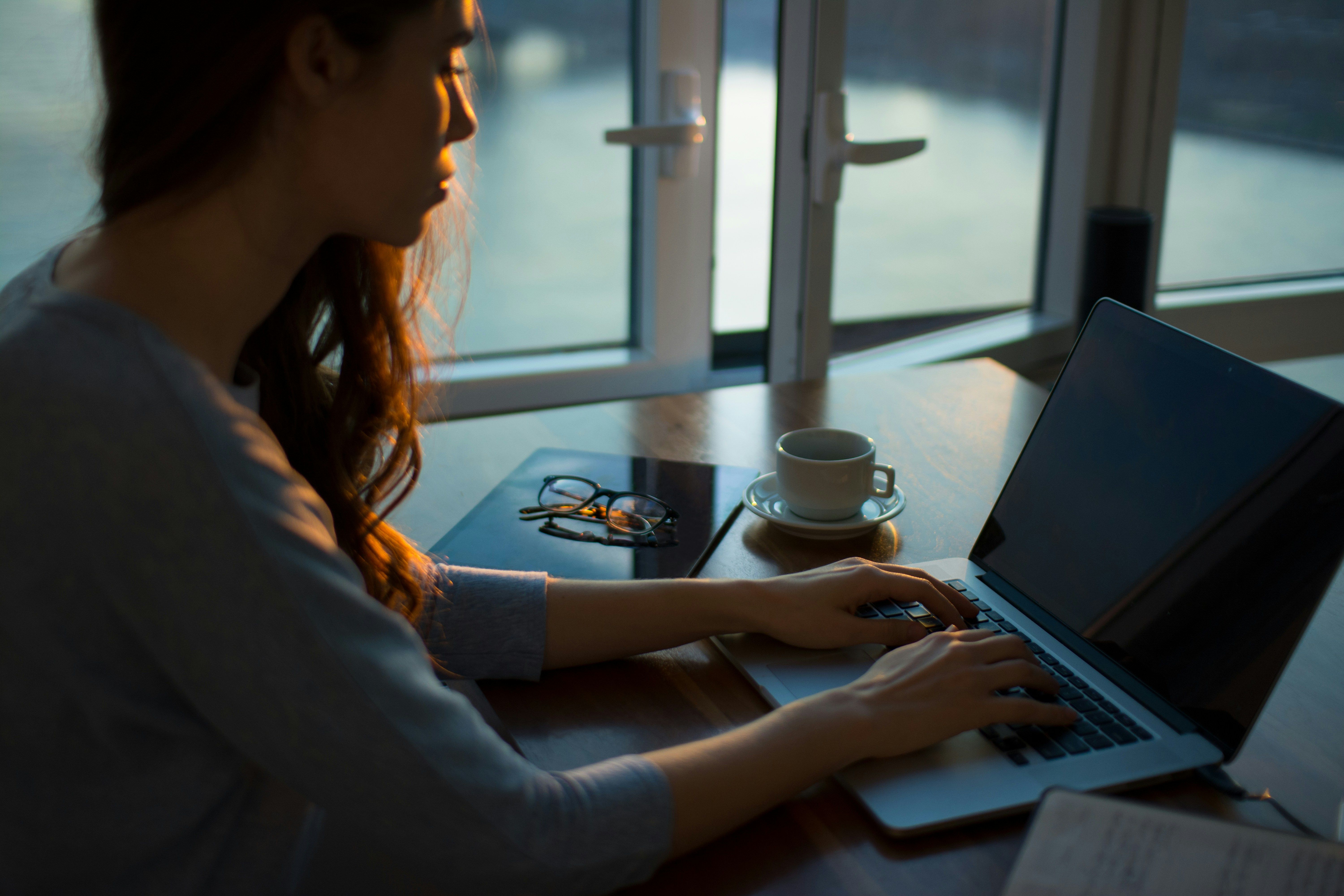 A woman with long red hair works on a laptop at a wooden table near a window at dusk, with a coffee cup and folded glasses beside her.