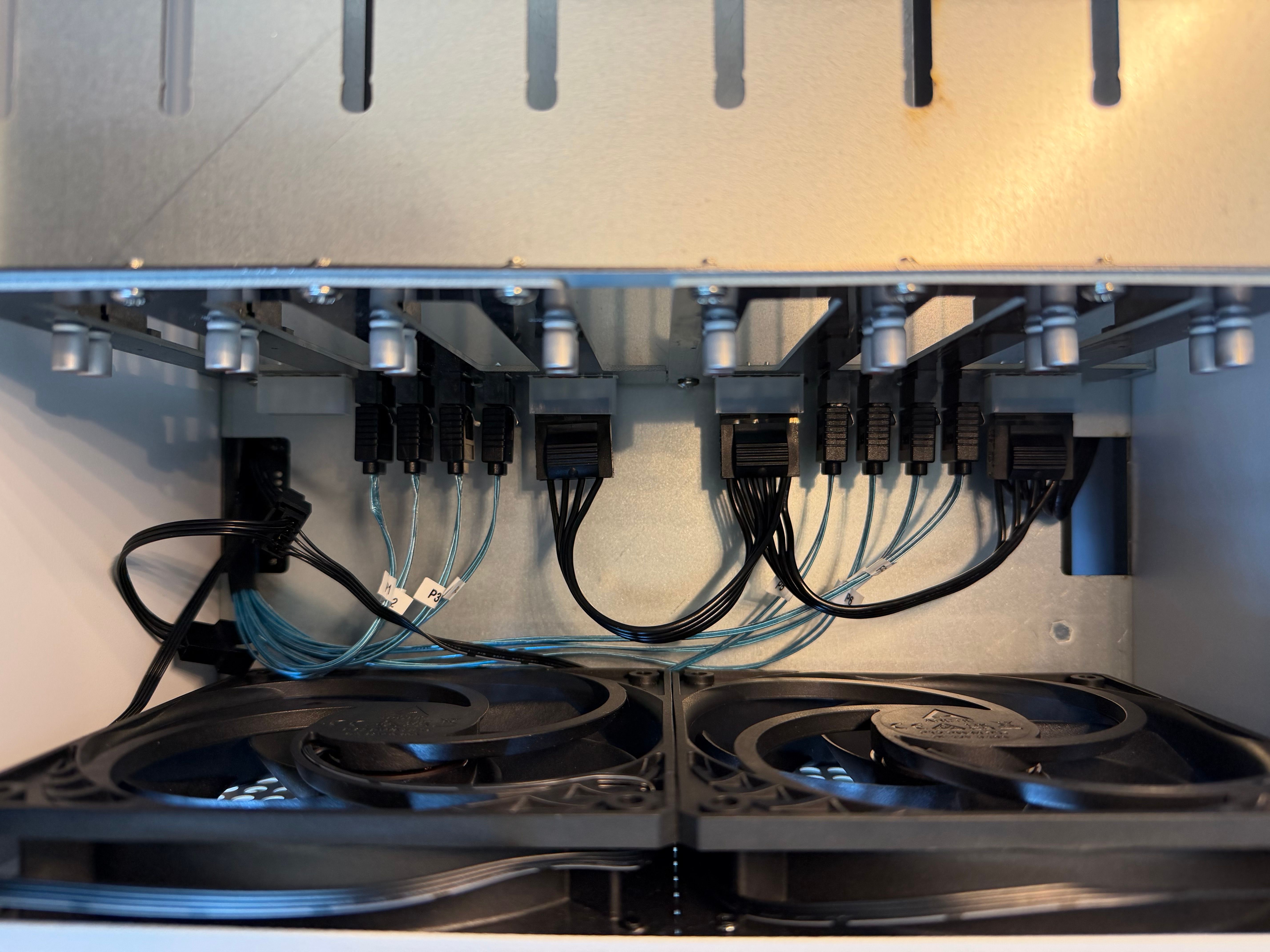 Interior of the NAS case showing labeled blue SATA cables and black power cables connected behind the eight drive bays, with two case fans below.