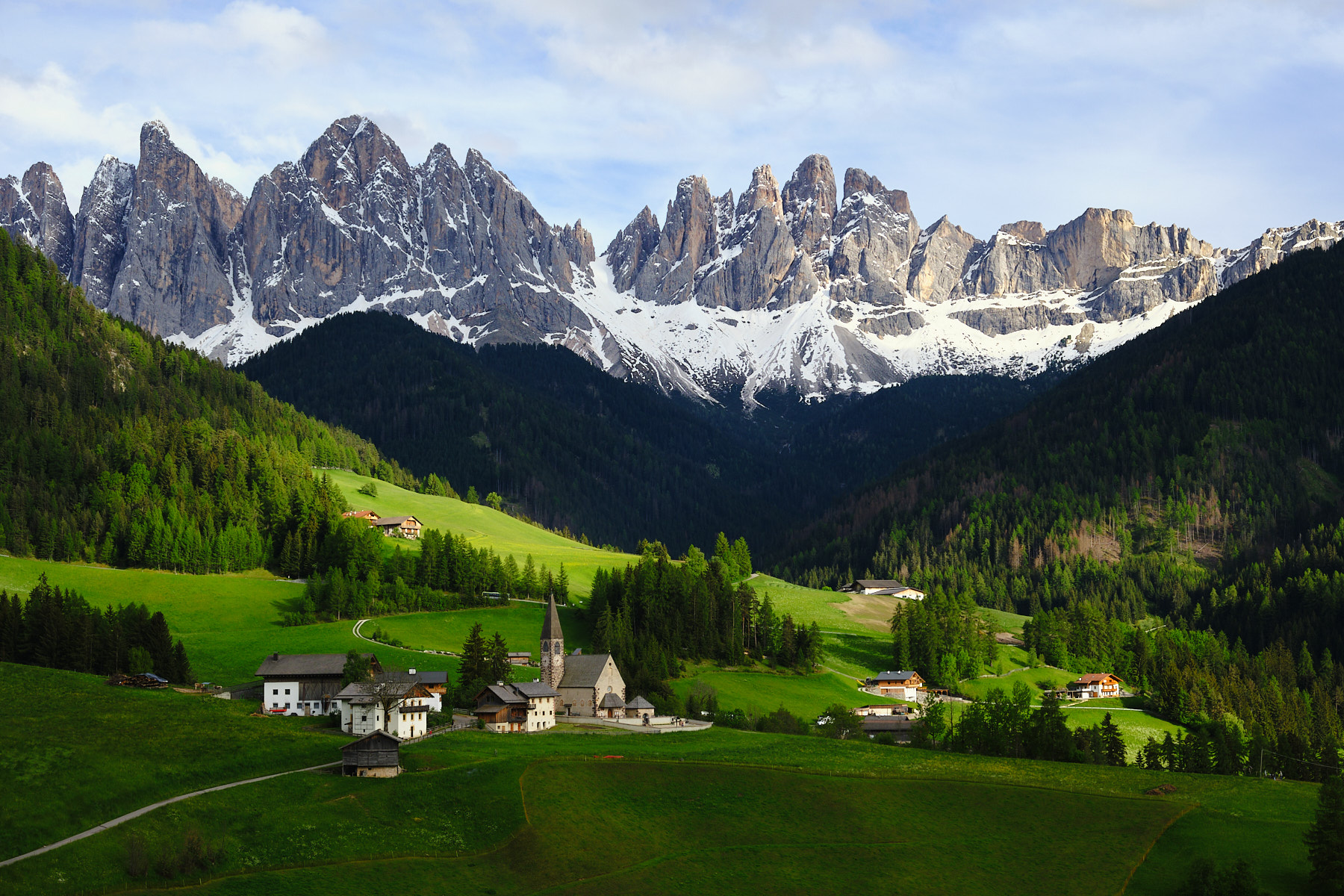 Alpine village with church in green valley beneath jagged Dolomite mountains.